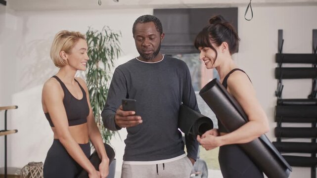 Male yoga instructor showing phone screen to female students. Women and yoga coach resting after yoga session. African American yoga teacher communicating with two women.