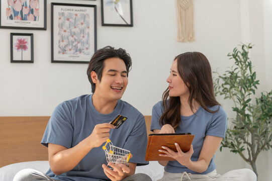 Young Asian couple shopping online while using digital tablet at home, Couple using credit card to pay bills or to book a room for vacation, making an online purchase.