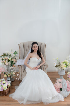 Wedding, Bride in beautiful dress sitting on sofa indoors in white studio interior, Young attractive Asian woman in wedding dress.