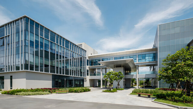 Modern glass office building with a connecting bridge and entrance canopy, surrounded by green trees and landscaping under a clear blue sky