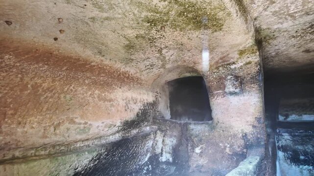 Salina, Malta - the curved interior of an ancient Paleo-Christian catacomb reveals a rock-cut passage with a dark rectangular opening, showcasing centuries of weathering, moss growth, and layered ston