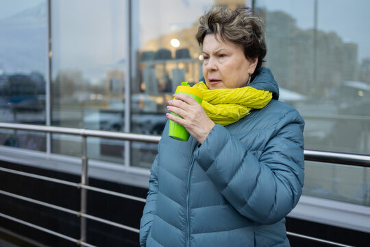Senior woman standing outdoors with takeaway mug, cold season clothing, modern city architecture behind, lifestyle concept.