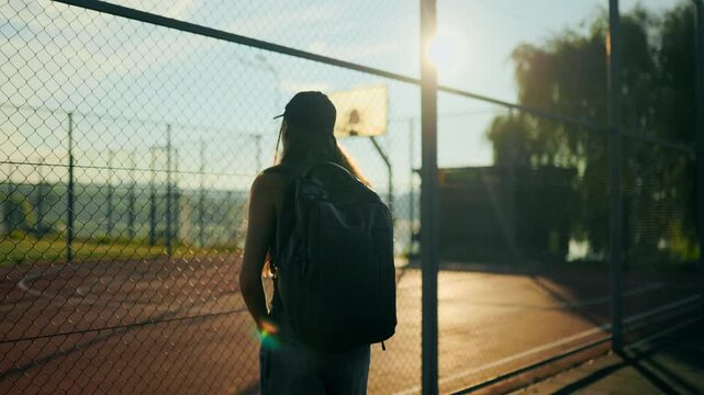 Back view of lonely preteen walking and stopping at sports fence.