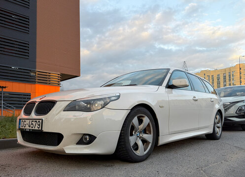 Front view of a white BMW 5 Series E61 station wagon car with an M Sport packet and black kidney grilles, parked outdoors near a modern building.
