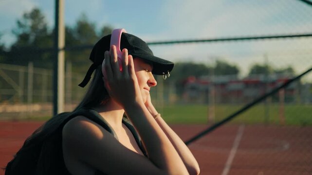Calm preteen girl putting on pink headphones near sports court at sunset.