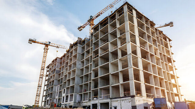 Tall concrete building under construction with two yellow tower cranes against a clear blue sky during a sunny day at the building site