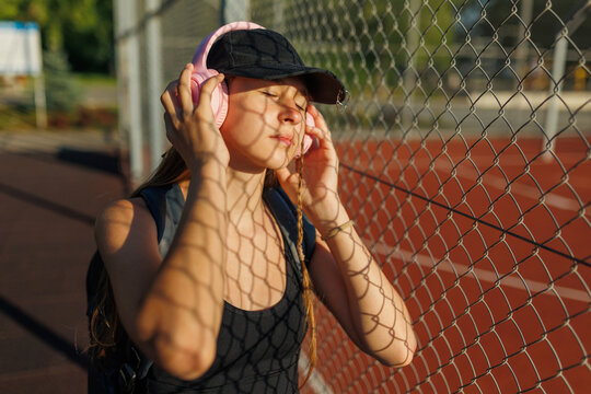Teenage girl with pink headphones resting by a sports fence during golden hour.