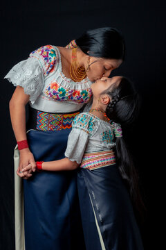 Indigenous Kichwa mother and daughter from Otavalo, Ecuador, wearing traditional hand-embroidered blouses and anaco sharing affectionate moments of cultural heritage in a studio portrait