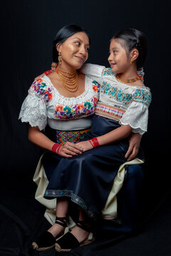 Indigenous Kichwa mother and daughter from Otavalo, Ecuador, wearing traditional hand-embroidered blouses and anaco sharing affectionate moments of cultural heritage in a studio portrait