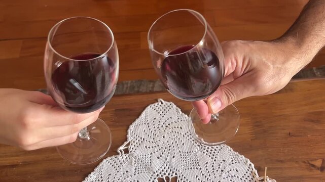 Romantic couple making a toast clinking glasses filled with red wine during a celebration, anniversary, or dinner over a wooden table with a classic doily