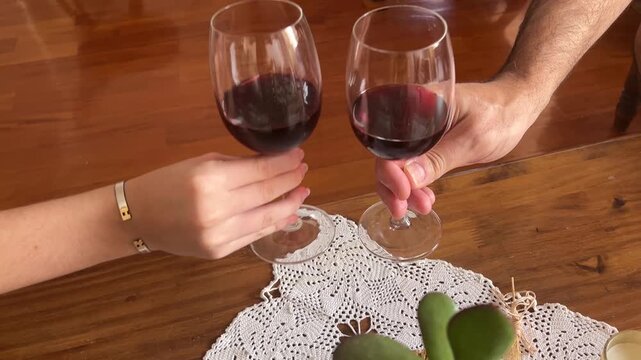 High angle view of a young woman and a man's hands holding glasses of red wine and making a toast over a rustic wooden table with a lace doily