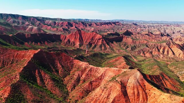 Aerial view of striking red sandstone formations in the Xinjiang Danxia landform, showing dramatic mountain ridges under a clear blue sky.