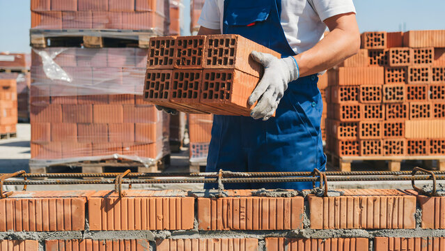 Construction worker wearing blue overalls and gloves holding a stack of red hollow bricks at a building site with pallets of bricks behind