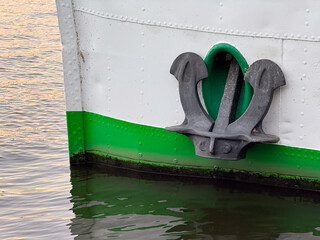 Boat anchor mounted on the bow of a vessel with a green stripe, partially submerged in calm water reflecting the surrounding environment © Vlad Yakubovskiy