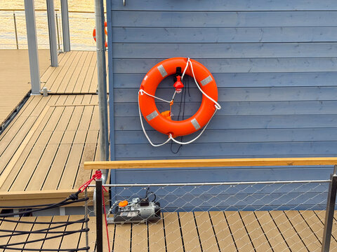 Orange life preserver mounted on blue wooden wall near wooden deck with safety equipment and railing visible in the background