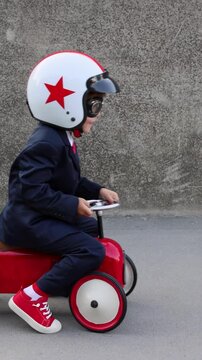 A happy child pretends to be a businessman. Funny kid rides a toy car outdoors. A boy has fun during the summer holidays. Back to school, the concept of a business idea. Slow motion, vertical video