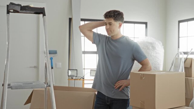 Man with hand on neck and hand on hip stands amid stacked cardboard boxes, ladder and bubble wrap in a bright moving room in building; tired contemplation.