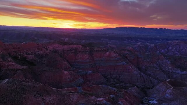Sunset casting vibrant orange and purple light over the rugged, multi-colored ridges of the Danxia landform in Xinjiang, China.