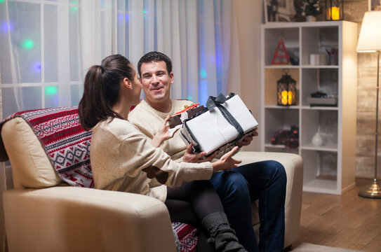 Young wife gives her husband a large Christmas present, showing a happy couple sharing a warm holiday celebration and gift exchange at home.