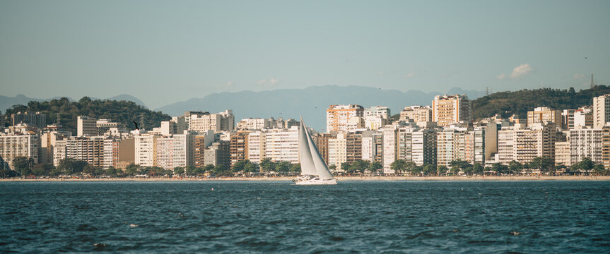 Compressed coastal cityscape panorama with a white sailing yacht gliding across blue bay water, dense beachfront apartment towers and layered mountains in clear daylight