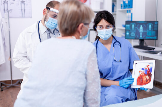 Senior woman discusses arrhythmia symptoms with a cardiologist during a masked hospital examination focused on diagnosis and care.