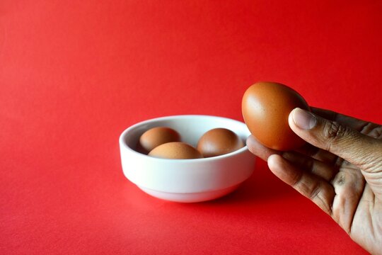 Close up of a hand holding a fresh brown egg next to a white ceramic bowl filled with eggs over a solid red background.