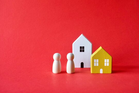 Two minimalist wooden peg figures standing next to a white and a yellow toy house model on a solid red background.