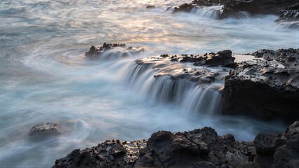 Fototapeta premium Ocean waves cascade over dark, rugged rocks in a serene long-exposure shot.