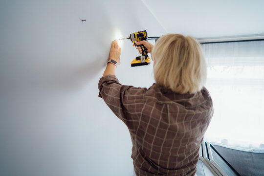 Back view middle aged woman in plaid shirt screwing a screw into the wall with electrical screwdriver. Woman wants to put a picture on the wall. Housekeeping work. Doing repair herself. DIY concept
