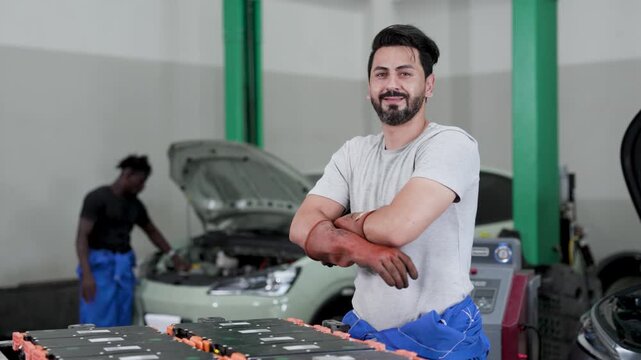 Auto technicians in a workshop servicing electric vehicles, with a large battery pack on a table and open hoods. Mechanic in blue overalls smiling, colleague working in background.