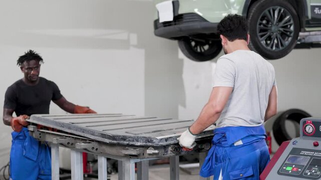 Two mechanics handle an electric car battery module beneath a lifted vehicle in a garage, performing EV maintenance with tools and safety gear.