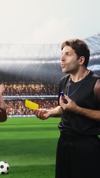 Vertical composition: Soccer referee showing yellow card to angry player protesting during match, blue teammates complaining about match official decision, disciplinary action sanctioning.