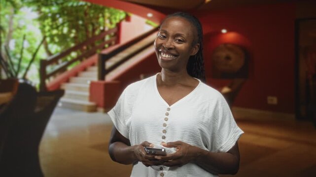 Woman holding smartphone with both hands, smiling broadly and looking at camera in a hotel lobby with staircase and plants; friendly joy.