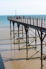 Fototapeta premium Bognor Regis West Sussex England UK. 14.07.2026. Two individuals relax on a pier structure with a weathered appearance, while another person is visible on the sandy beach below