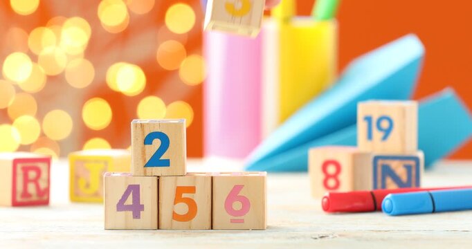 Woman putting cubes with numbers, letters, stationery and paper plane at wooden table against orange background with blurred lights, closeup. Bokeh effect