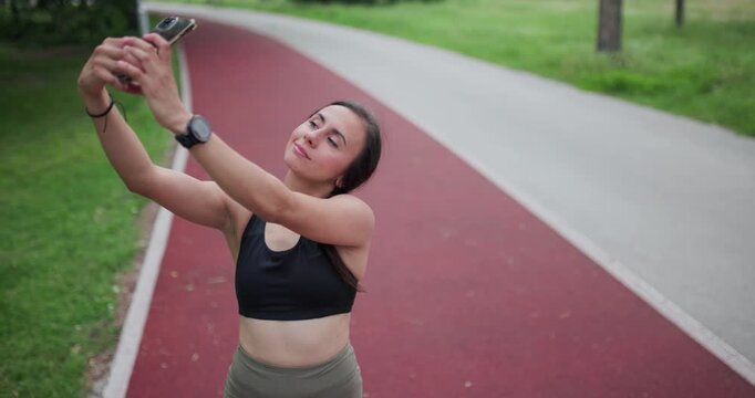 A Young Woman in Sportswear Takes a Selfie with Her Smartphone on a Red Running Track in a Lush Green Park, Capturing Her Fitness Journey and Healthy Lifestyle Moment.