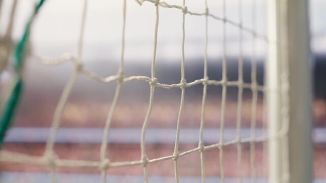 Selective focus on goal net and post detail on football pitch under stadium light, with side-post rope knots, blurred soccer field, and sports equipment in the foreground.