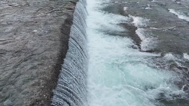 Powerful waterfall cascades over rugged stone edge. Foam churns as water crashes into turbulent downstream river. Natural force dominates the scene with raw, untamed energy