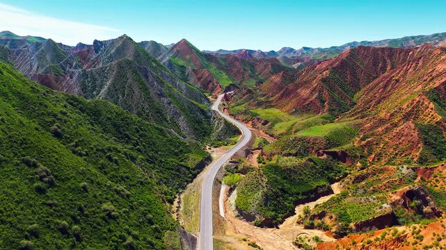 Aerial view of winding asphalt highway cutting through a dramatic valley with lush green hills on one side and vibrant red rock formations on the other under a clear blue sky.