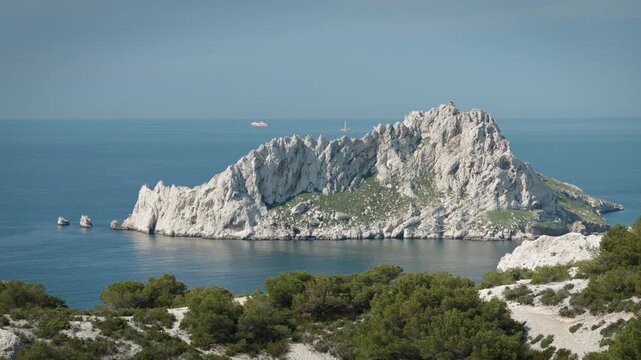 A large, steep island rising out of the deep-blue water in the Calanques National Park in France. The chalk cliffs glisten in the low evening sun. Sailboats are out on the water.