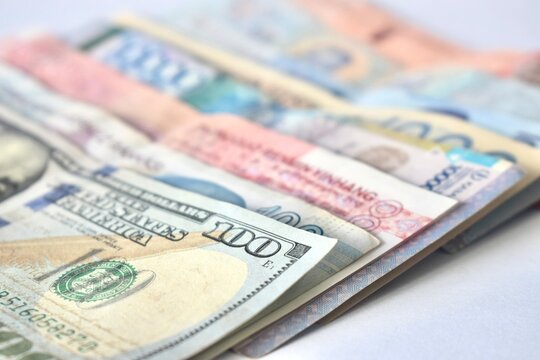 A low-angle, selective focus close-up shot of a United States hundred-dollar bill layered over multiple international currency banknotes on a white surface.