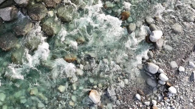 Clear water flows over smooth stones. Currents swirl, creating frothy white patterns. Rocks vary in size, color, and texture. Shallow stream reflects natural movement