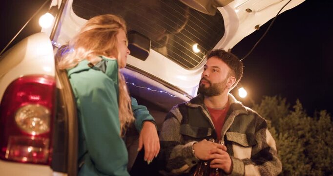 Couple talks near car at night with string lights hanging above in a relaxed outdoor setting by trees