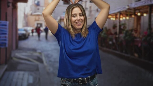 Woman making a heart with hands above head, smiling with visible forearms, standing on a cobbled street outside a cafe; joy affection travel.