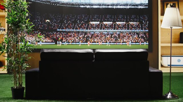 Empty living room with football game on television broadcast and couch facing large display, with a home entertainment setup made of TV, cozy sofa and relaxed match night setting.