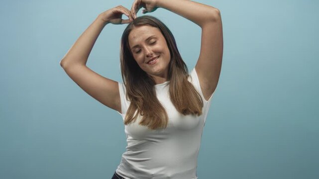 Woman forming heart with hands above head, white tshirt exposing bare forearms, smiling in studio with blue seamless backdrop; playful affection.