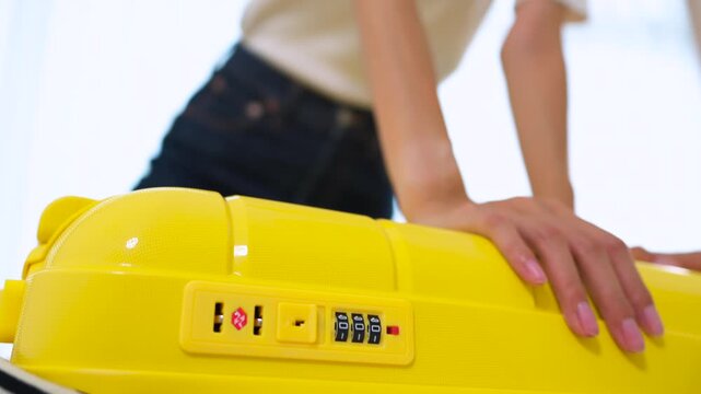 Close-up of Asian woman sitting on an overpacked yellow suitcase to close it. Concept of overpacking issues, travel preparation struggles and holiday planning.