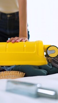 Close-up of Asian woman sitting on an overpacked yellow suitcase to close it. Concept of overpacking issues, travel preparation struggles and holiday planning. Vertical Shot