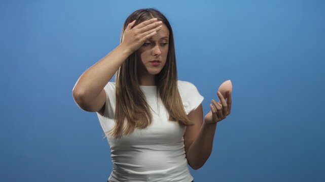 Woman holding a pink makeup sponge while pressing hand to forehead against a blue studio backdrop, wearing white tee and long brunette hair; frustration beauty doubt.