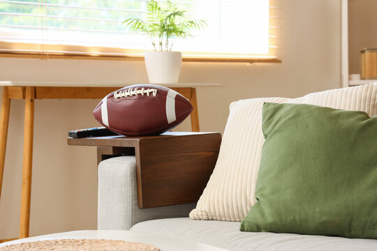 American football and remote control on a wooden sofa armrest tray in a cozy living room setting, the cozy living room features a light-colored sofa with beige and green cushions, a window